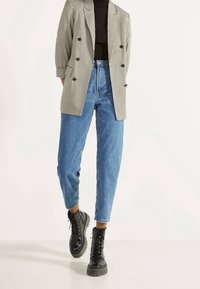Blazer doppiopetto in bianco e nero a scacchi houndstooth con grandi bottoni, indossato sopra un dolcevita nero e jeans a vita alta blu; stivali neri con lacci.