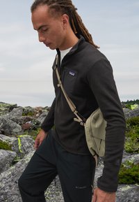 Felpa nera con zip e motivo testurizzato, abbinata a pantaloni neri e a una borsa a tracolla beige. Ambiente esterno con terreno roccioso e vegetazione.