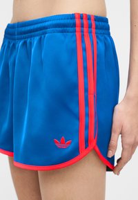 Μπλε αθλητικό σορτς με κόκκινο τελείωμα και τρεις κάθετες κόκκινες ρίγες, με κόκκινο λογότυπο Adidas κοντά στο τελείωμα στο γοφό ενός ατόμου.