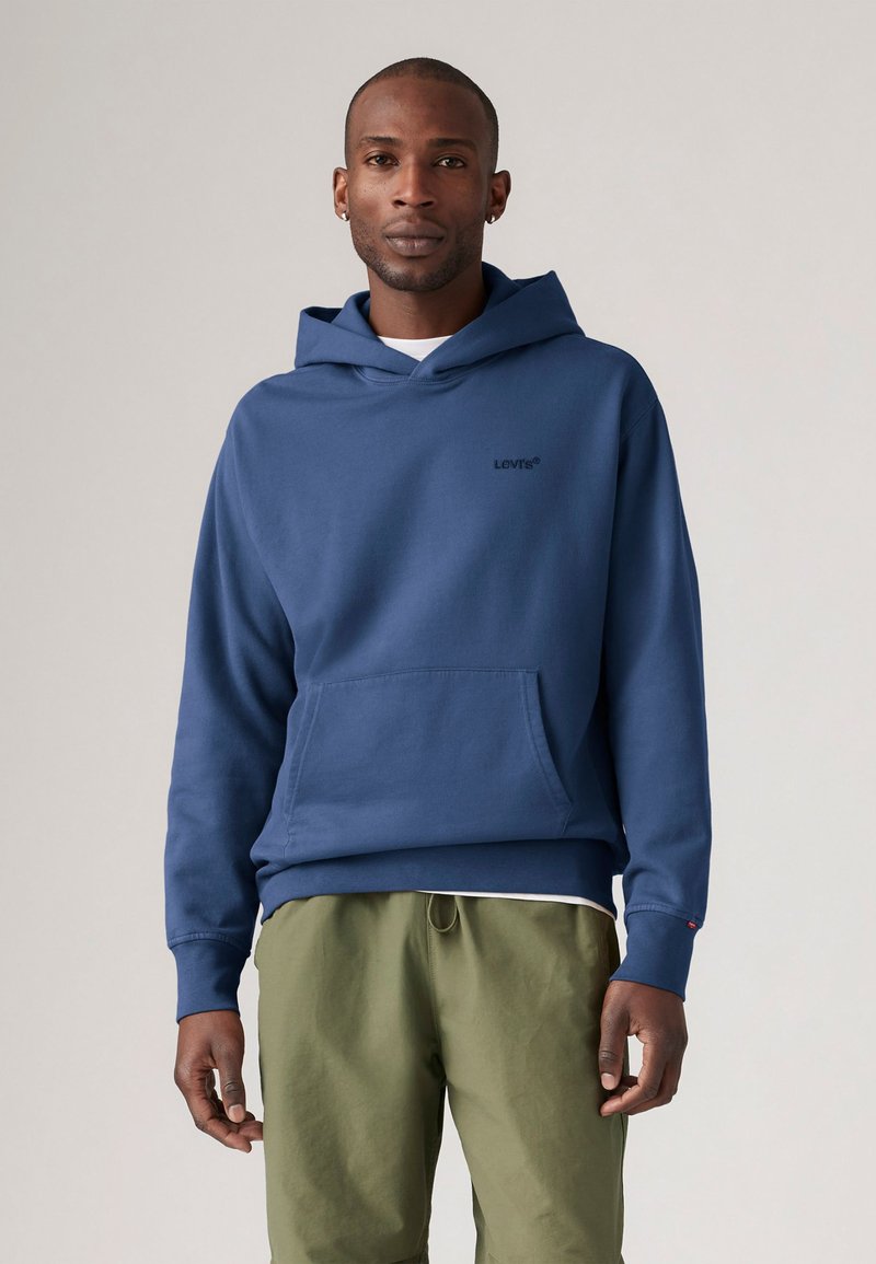 Námornícky modrá mikina s kapucňou a prednou kapsou, vyrobená z bavlneného materiálu, s minimalistickým logom Levi's. V kombinácii s olivovozelenými nohavicami.