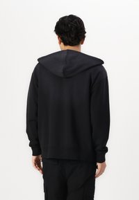 Črna kapucinska majica z ohlapnim krojem, rebrastimi manšeti in hemom ter mehkim tekstilom, prikazana s hrbtne strani.