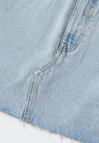 Tela de denim de color azul claro con una superficie texturizada, con costuras amarillas a lo largo de las costuras y bordes deshilachados en la parte inferior.