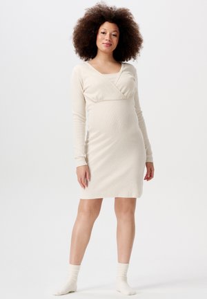 Beige Schwangerschafts-Kleid mit langen Ärmeln, mit überkreuztem Ausschnitt, figurbetontem Schnitt und weicher Textur, kombiniert mit weißen kniehohen Socken.