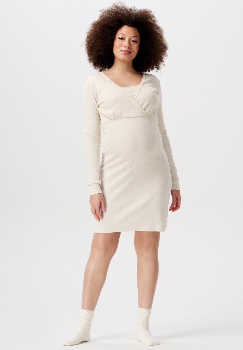 Beżowa sukienka ciążowa z długim rękawem, z dekoltem w kształcie krzyża, dopasowana sylwetka i miękka tekstura, zestawiona z białymi skarpetkami do kolan.