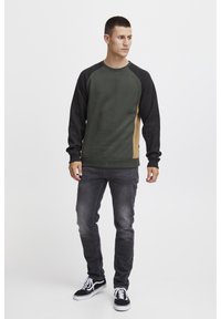 Mørkegrøn sweatshirt med sorte ærmer og beige detaljer, med rund halsudskæring og ribbede manchetter, parret med slidte sorte jeans og sneakers.