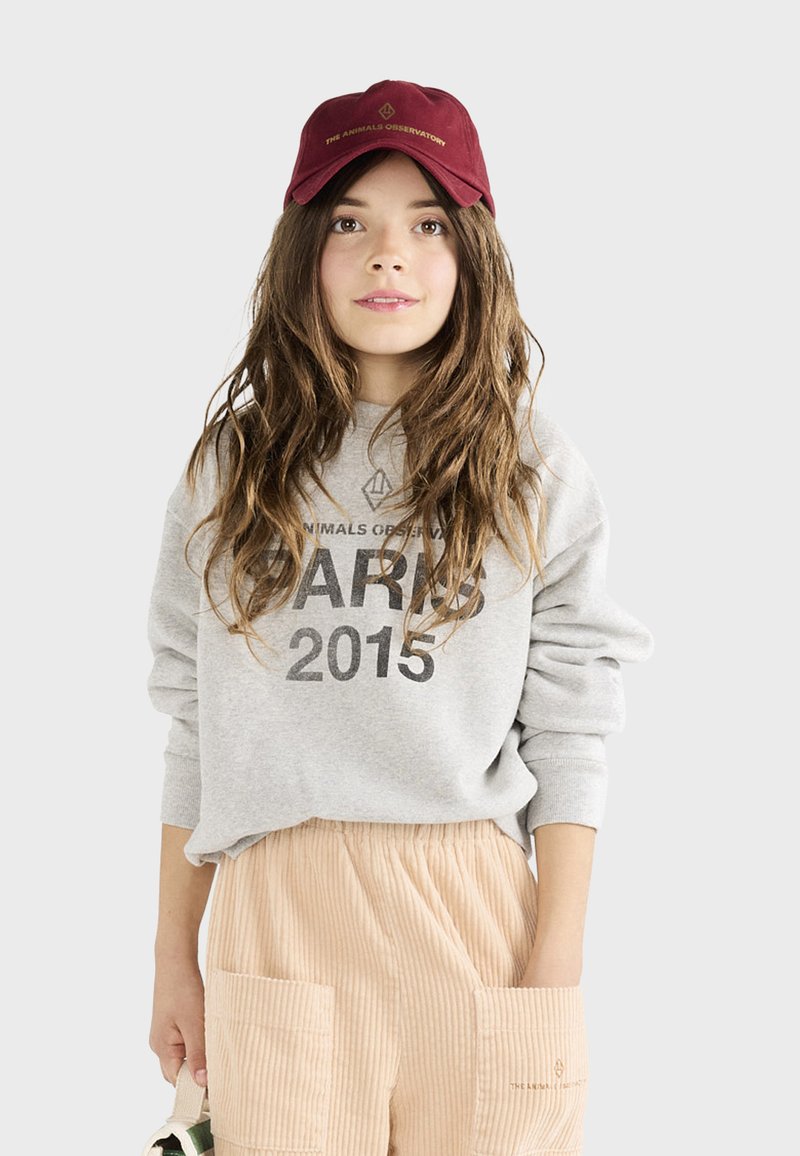 Sudadera gris con estampado de "PARÍS 2015", combinada con pantalones de pana de color beige claro que cuentan con bolsillos. Gorra burdeos con acentos de logo.