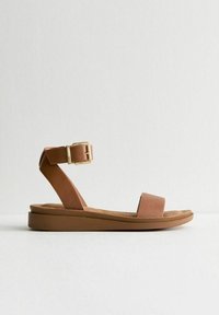 Sandales plates en cuir marron avec une large sangle sur les orteils, une sangle à la cheville et un accent de boucle dorée. La semelle est plate et rembourrée.