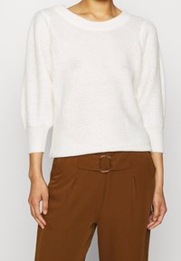 Pull blanc en tricot avec un col rond et des manches larges, associé à un pantalon marron avec un détail de ceinture circulaire.