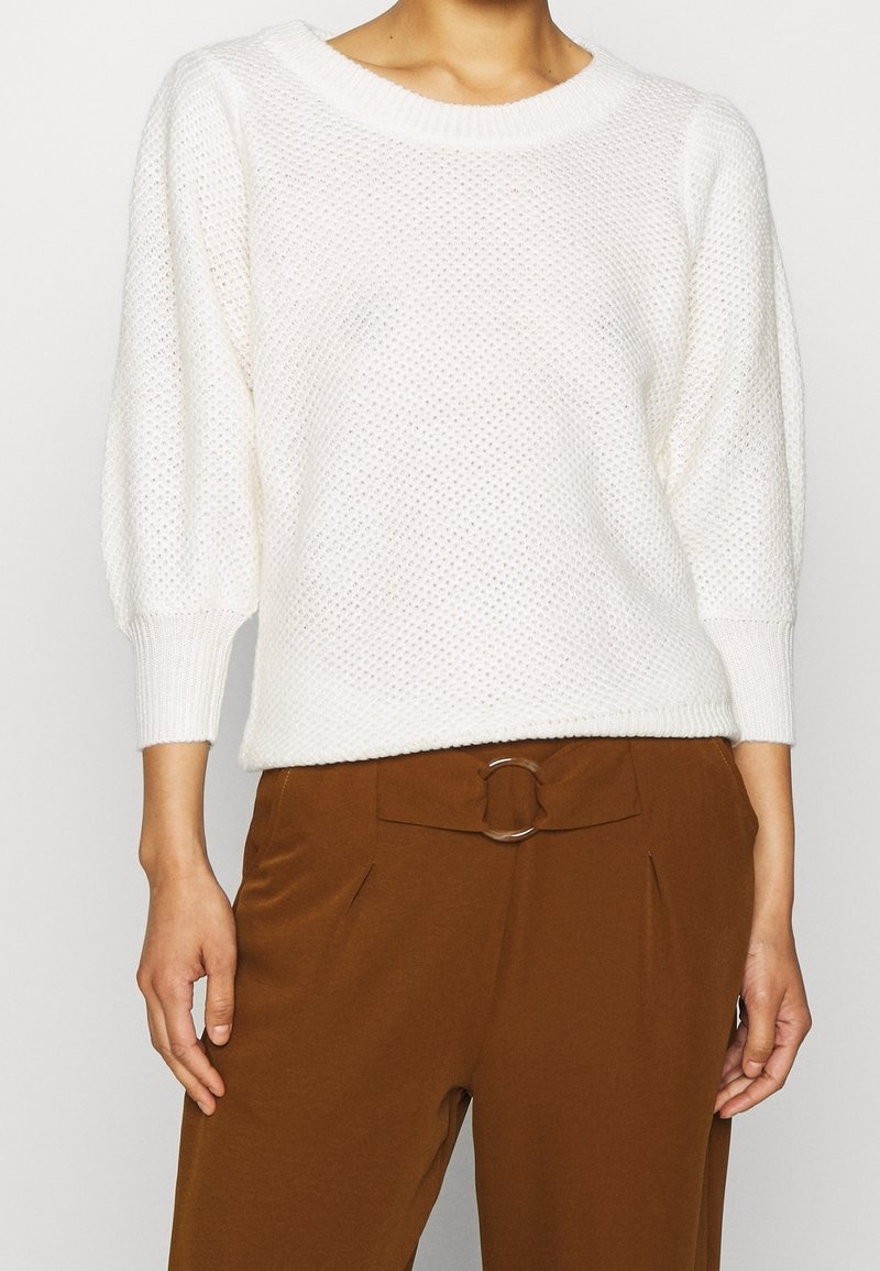 Pull blanc en tricot avec un col rond et des manches larges, associé à un pantalon marron avec un détail de ceinture circulaire.