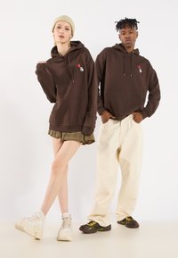 Bruine hoodies met een voorzak en geborduurde ontwerpen, gecombineerd met beige baggy broeken en sneakers. Model draagt een geruite rok.