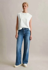 Witte mouwloze top met gestructureerde schouders, gecombineerd met lichtblauwe wijde jeans. Het model draagt een crèmekleurige sneakers en een eenvoudige ketting.