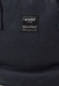 Svart kanvasryggsäck med rektangulär form, texturerad yta och en synlig etikett med varumärke och designinformation.