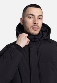 Chaqueta aislante negra con cuello alto y capucha. Presenta una cremallera frontal y puños elásticos. Tejido suave con un acabado mate.