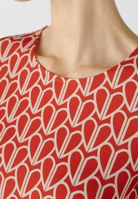 Un haut en tissu rouge présente un motif de cœurs répété en crème, avec un col rond et une texture lisse. Les détails des coutures sont visibles.