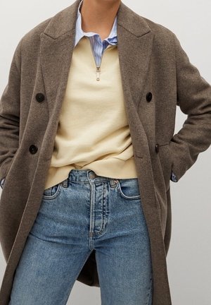 Bruine dubbelzijdige jas over een beige ritssweater, gecombineerd met lichtblauwe jeans. De jas heeft zwarte knopen en gestructureerde schouders.