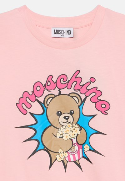 Розова детска тениска с меченце, държащо пуканки, и думата "moschino" с розови букви над него на син фон с лъчи.