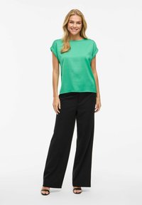 Top in satin verde con maniche corte, abbinato a pantaloni neri a vita alta e gamba larga, insieme a sandali neri con fascia. Tessuto liscio, vestibilità rilassata.
