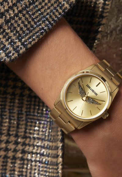 Zlaté náramkové hodinky s lesklým povrchom, s detailom krídla na ciferníku, pripojené na zlatý náramok s článkami v rovnakom prevedení.