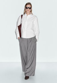 Femme marchant avec une chemise blanche oversized, un pantalon gris large, des lunettes de soleil noires, portant un sac à épaule marron, sur un fond blanc uni.