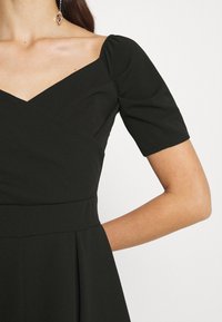 Femme portant une robe noire avec un décolleté en cœur, des manches courtes bouffantes et une taille cintrée sur un fond blanc.
