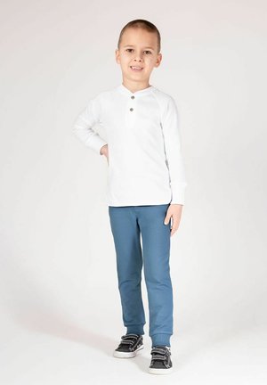 Biała koszula z długim rękawem, zapinana na guziki przy kołnierzu, zestawiona z niebieskimi spodniami i czarnymi trampkami. Tkanina wygląda na gładką i casualową.