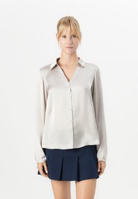 Blonde vrouw met een pony, gekleed in een lichtbeige satijnen blouse met knopen en een marineblauwe geplooide minirok, staand tegen een witte achtergrond.