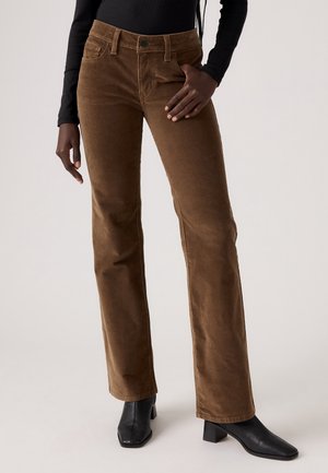 Vrouw met bruine corduroy bootcutbroek, zwart shirt met lange mouwen en zwarte enkellaarsjes met hak, staand met één hand in haar zak.