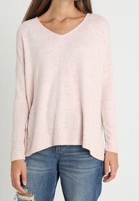 Ljusrosa v-ringad tröja med långa ärmar, lös passform och textilstruktur. Matchad med slitna blå jeans.