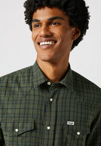 Chemise à carreaux verts avec des lignes navy, col boutonné et deux poches poitrine. Présente des boutons blancs et une petite étiquette de marque sur le devant.