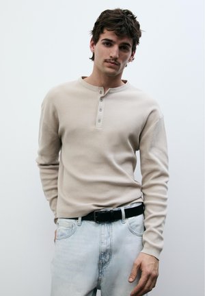 Junger Mann mit Schnurrbart, der einen beigen Henley-Pullover und hellblaue Jeans mit schwarzem Gürtel trägt und vor einem weißen Hintergrund steht.