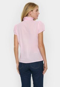 Kortärmad rosa blus med en volangkrage, tillverkad av lättviktsmaterial. Har en avslappnad passform och slät textur. Bärs med mörka denimjeans.