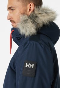 Parka azul marino con capucha forrada de piel sintética, cordón naranja y un parche de cuero negro con letras plateadas en relieve en la manga.