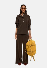 Brązowy oversize pulower z detalami w postaci guzików, w połączeniu z dopasowanymi szerokimi spodniami. Trzyma żółty zamszowy plecak z zamkami i paskami.