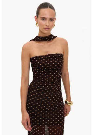 Femme aux cheveux tirés en arrière porte une robe noire sans bretelles à pois marron, des boucles d’oreilles créoles dorées et un bracelet épais en or sur fond blanc.