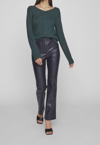 Pull col en V couleur sarcelle avec manches longues et poignets côtelés, associé à un pantalon en cuir noir ajusté et des sandales noires à talons.