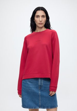 Kvinde med langt sort hår iført en rød langærmet sweatshirt og en blå denimnederdel, stående mod en ensfarvet hvid baggrund.