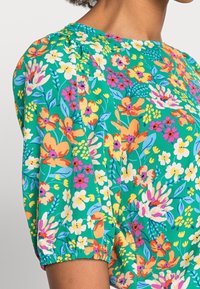 Haut à motifs floraux colorés avec un fond vert, des manches bouffantes et un col rond. Le tissu a une texture douce.