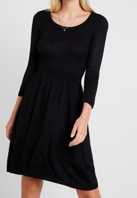 Robe noire en tricot avec un corsage côtelé, des manches trois-quarts et une jupe évasée. Texture lisse, taille cintrée et élasticité subtile.