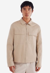 Beige colbert gemaakt van duurzame stof, met een ritssluiting aan de voorkant, twee borstzakken met kleppen en een rechte snit.