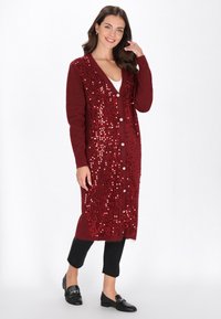 Femeie îmbrăcată într-un cardigan burgund până la genunchi, împodobit cu paiete, pantaloni negri și mocasini negri, zâmbitoare și atingându-și părul, pozând pe un fundal alb.