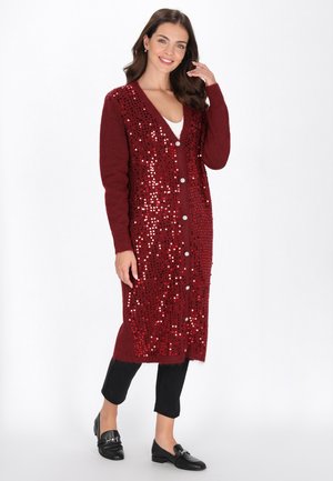 Femeie îmbrăcată într-un cardigan burgund până la genunchi, împodobit cu paiete, pantaloni negri și mocasini negri, zâmbitoare și atingându-și părul, pozând pe un fundal alb.