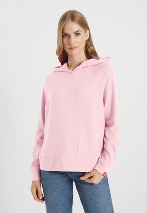 Jonge vrouw met een lichtroze capuchontrui met voorzakken en een blauwe spijkerbroek, staande tegen een effen lichte achtergrond.