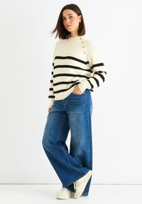 Cream sweater with black stripes, ribbed texture, and button accents on the shoulder, paired with wide-leg blue jeans and white sneakers.