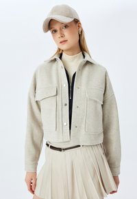Hellgraue, cropped Jacke mit zwei frontalen Taschen, silbernen Druckknöpfen und einem kragenlosen Hals, kombiniert mit einem beigen Faltenrock und einer Mütze.