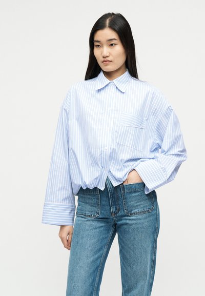 Γυναίκα με μακριά μαύρα μαλλιά που φοράει μεγάλη πουκαμίσα με γαλάζιες και λευκές ριγέ λεπτομέρειες και ψηλόμεσα μπλε τζιν, στέκεται με το ένα χέρι στην τσέπη.