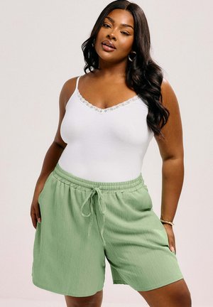 Frau mit langen dunklen Haaren, die ein weißes Tanktop mit Spitzenbesatz und hellgrüne Shorts mit Kordelzug trägt, stehend mit den Händen in den Taschen.