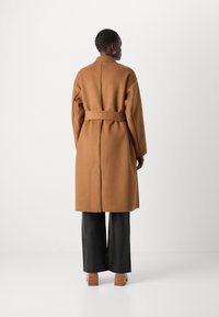 Brauner Wollmantel mit tailliertem Gürtel, weiten Ärmeln und geradem Schnitt. Kombiniert mit schwarzen Hose mit weiter Beinform und Holzabsatzschuhen.