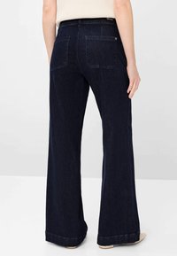 Persona che indossa jeans larghi blu scuro con tasche posteriori, in piedi con una mano rilassata lungo il fianco contro uno sfondo semplice.