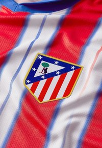 Punainen ja valkoinen raidoitettu jalkapallopaita, jossa on tähdillä varustettu vaakuna ja puumuotoilu. Kankaan pinta on tekstuuri.