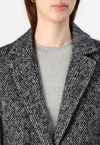 Manteau en motif pied-de-poule noir et blanc avec un col cranté, superposé sur un haut en jersey gris clair à côtes. Texture lisse avec un design structuré.